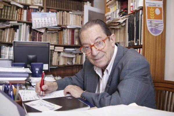 Hombre escribiendo en un escritorio rodeado de libros y documentos.