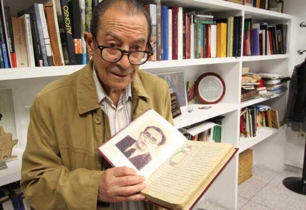 Herminio Ramos sosteniendo un libro con su retrato en una biblioteca