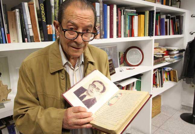 Herminio Ramos sosteniendo un libro con su retrato en una biblioteca