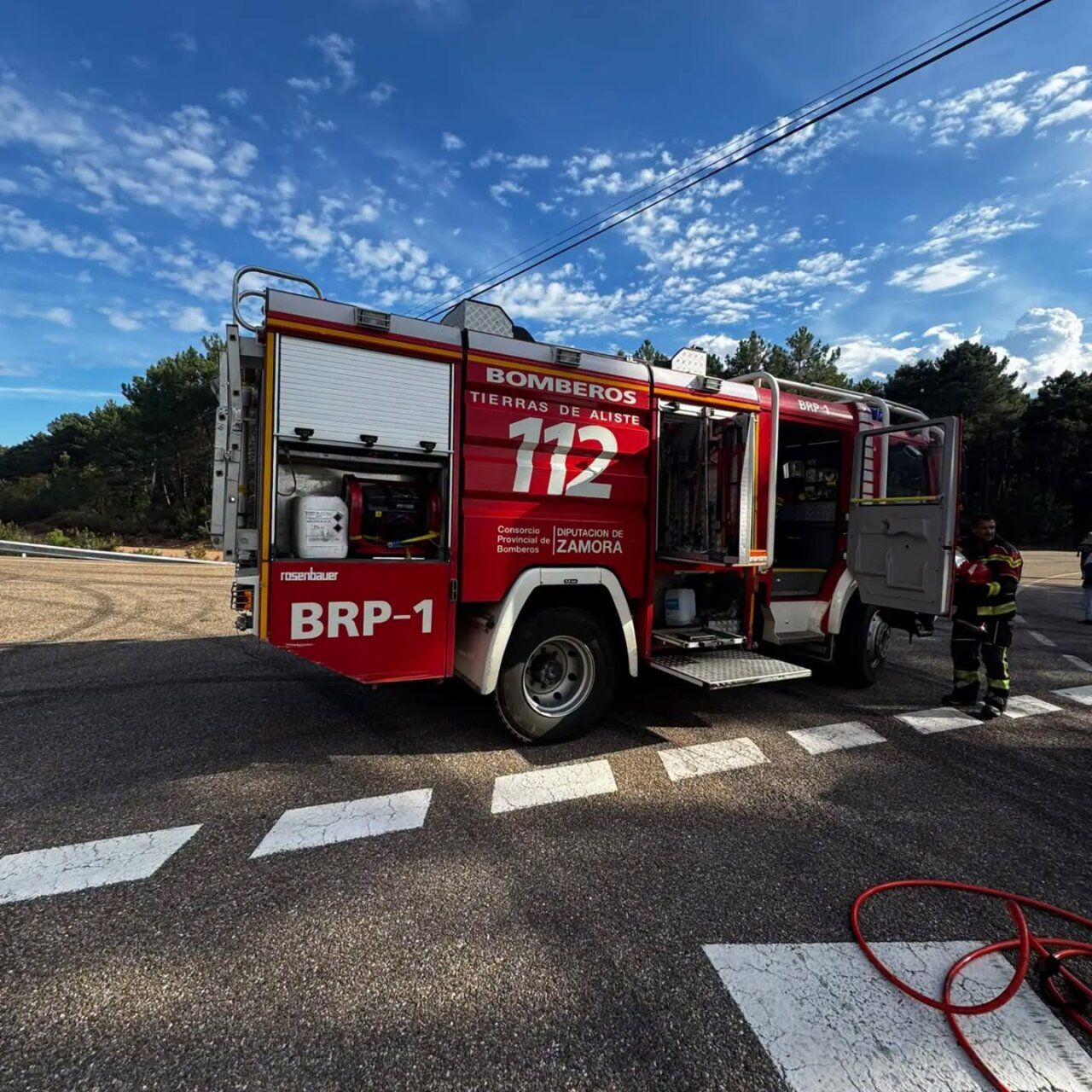 Camión rojo de bomberos estacionado en una carretera, con compartimentos abiertos y un bombero al lado.