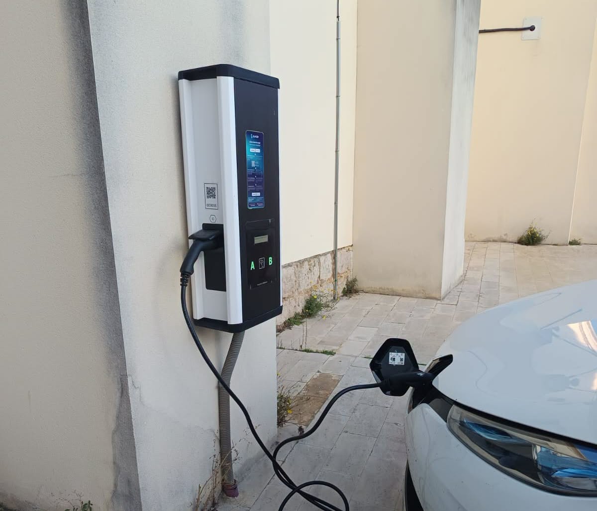 Un coche blanco conectado mediante cable a un punto de recarga junto a una pared.