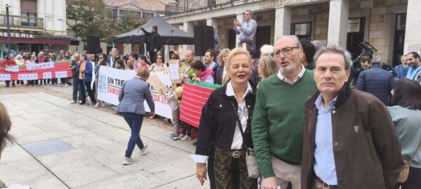Manifestantes sostienen pancartas sobre el tren en una plaza, junto a tres personas en primer plano.