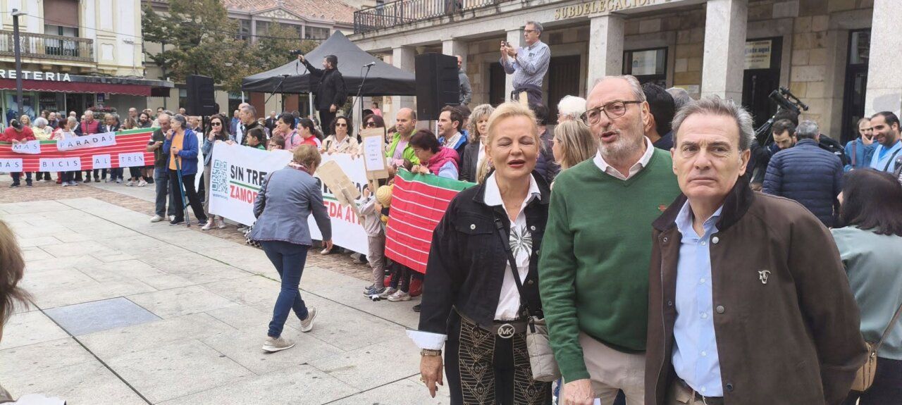 Manifestantes sostienen pancartas sobre el tren en una plaza, junto a tres personas en primer plano.