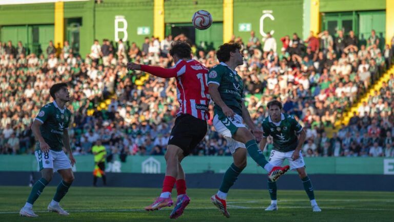 Futbolistas de rojo y verde disputan un balón aéreo ante las gradas