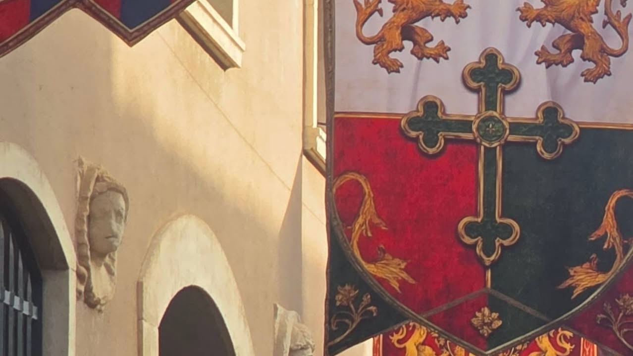 Detalle de un estandarte con cruz junto a una fachada de piedra con arcos.