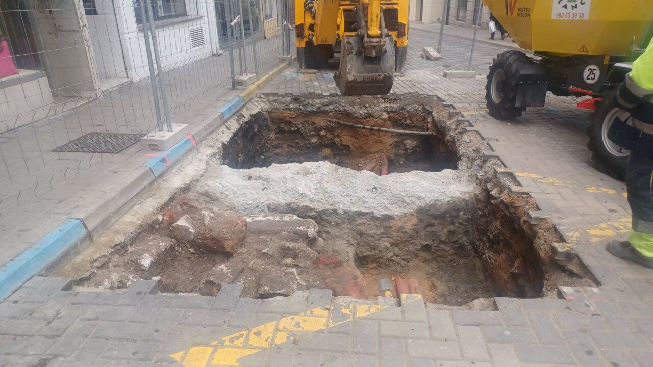 Excavación abierta en una calle, con una excavadora y vallas de obra alrededor.