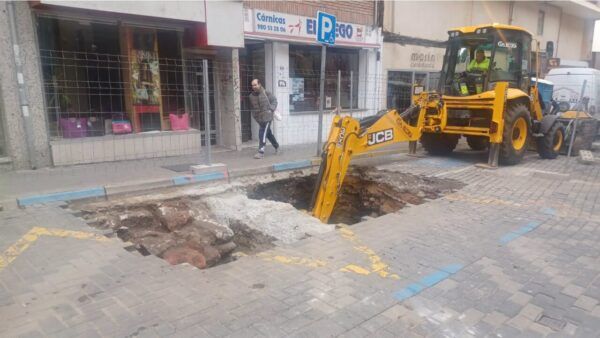 Una retroexcavadora trabaja junto a una excavación abierta en una calle adoquinada