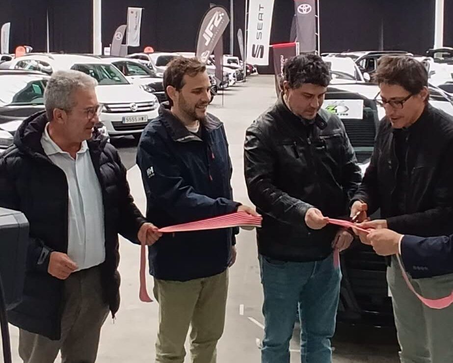 Varias personas sostienen una cinta roja entre coches en una exposición de automóviles.