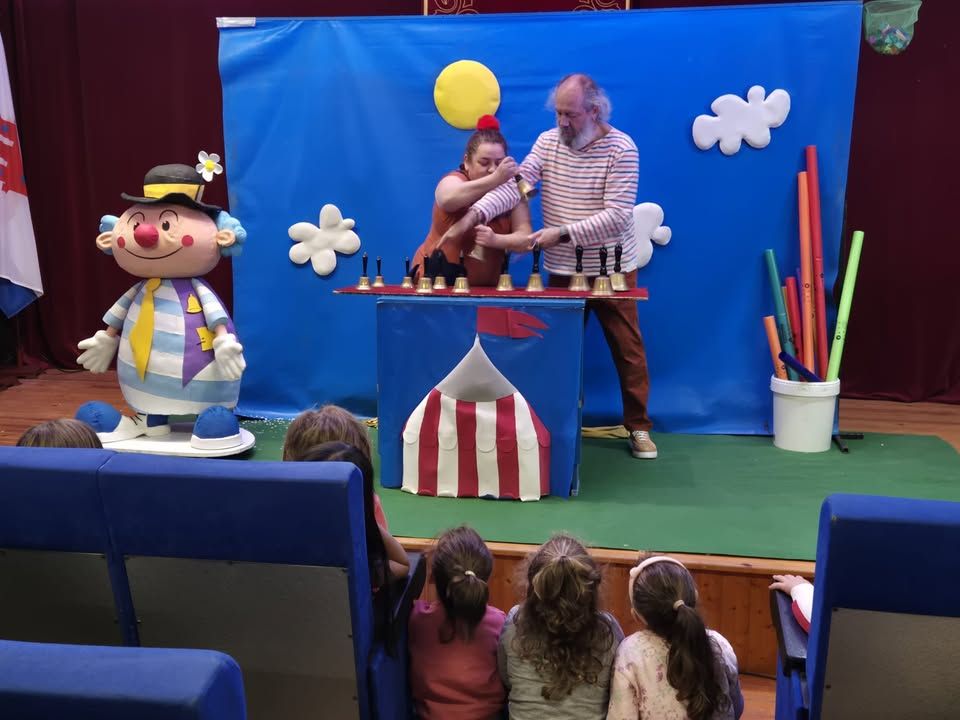 Dos personas actúan tras una mesa mientras varios niños observan desde sus asientos.