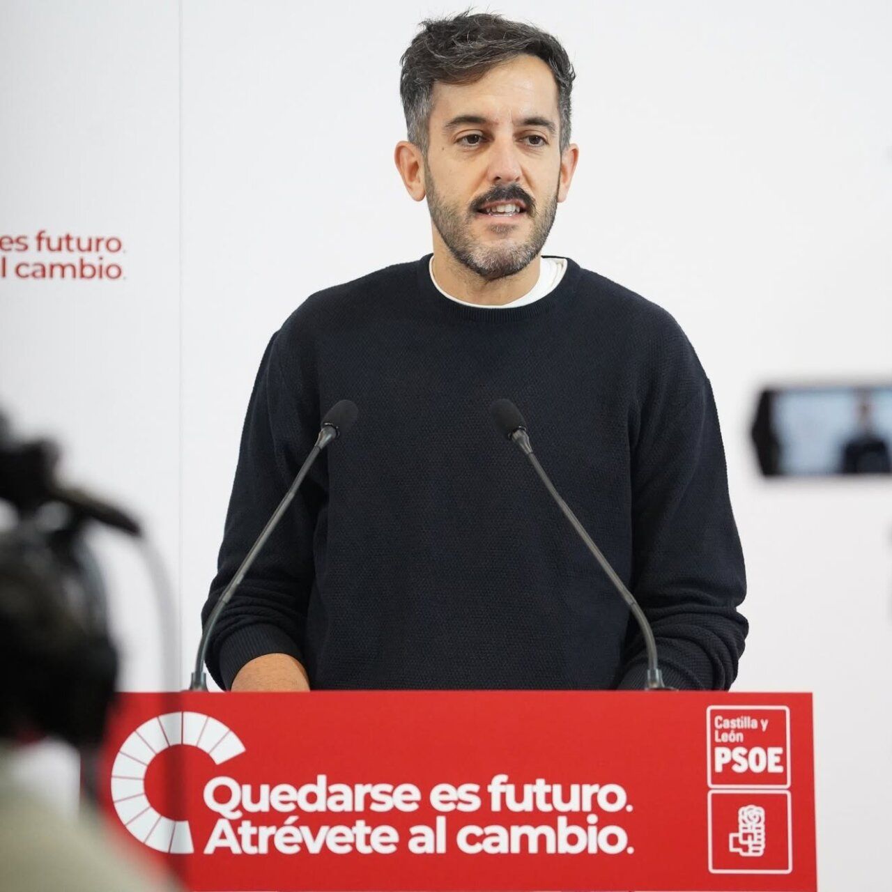 Hombre de pie ante un atril rojo del PSOE de Castilla y León con dos micrófonos.