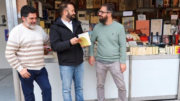 Tres hombres conversan frente a un puesto con libros; uno sostiene un ejemplar abierto.