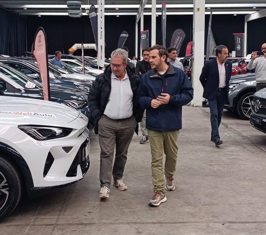 Dos hombres recorren una exposición interior con varios automóviles alineados.
