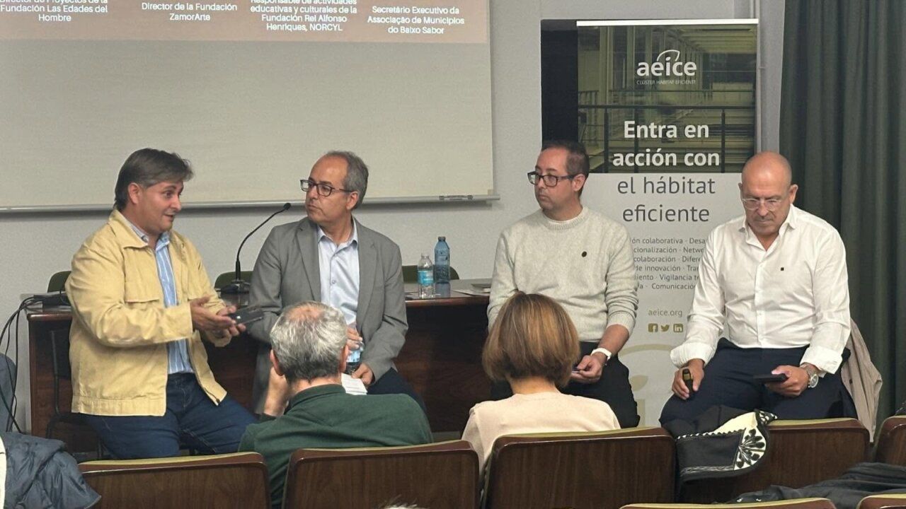 Cuatro hombres sentados frente a asistentes durante un encuentro en una sala con pantalla y cartel.