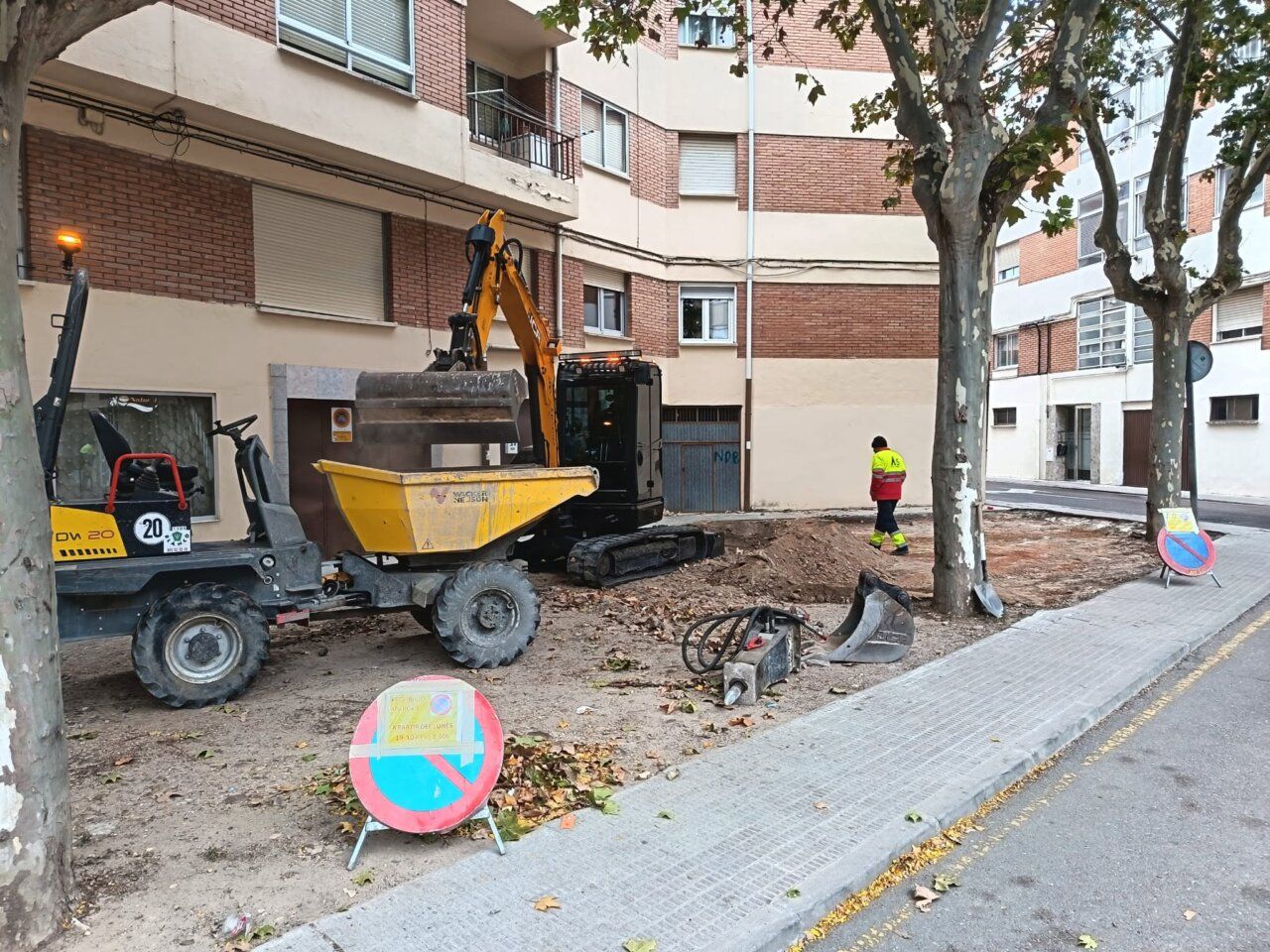 Maquinaria y un operario trabajan en un terreno junto a una acera arbolada