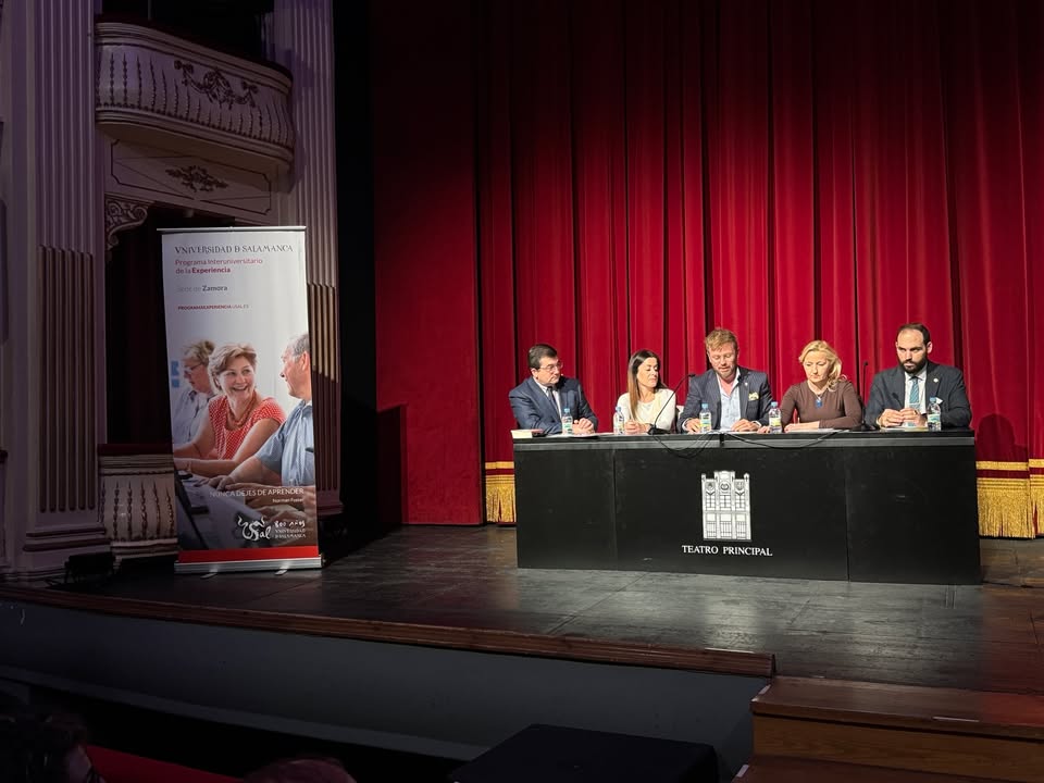 Cinco personas sentadas en una mesa sobre el escenario, ante un telón rojo.