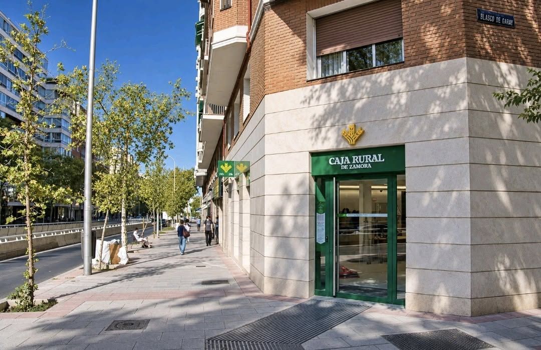Fachada de una oficina de Caja Rural de Zamora junto a una acera arbolada.
