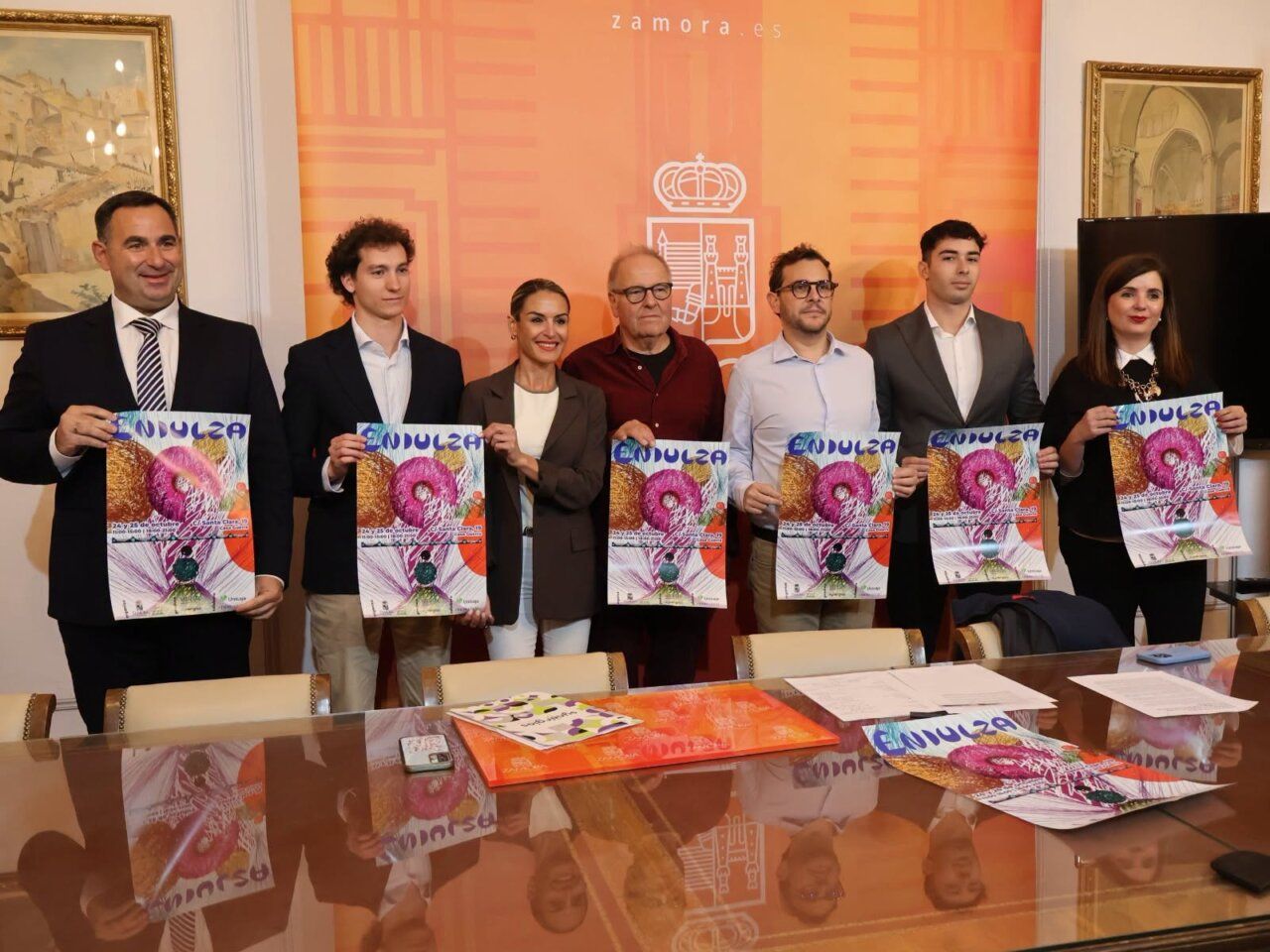 Siete personas sostienen carteles de ENDULZA frente a un fondo naranja con un escudo