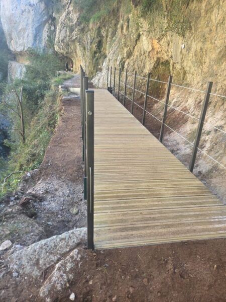 Pasarela de tablones con barandilla metálica junto a una pared rocosa.