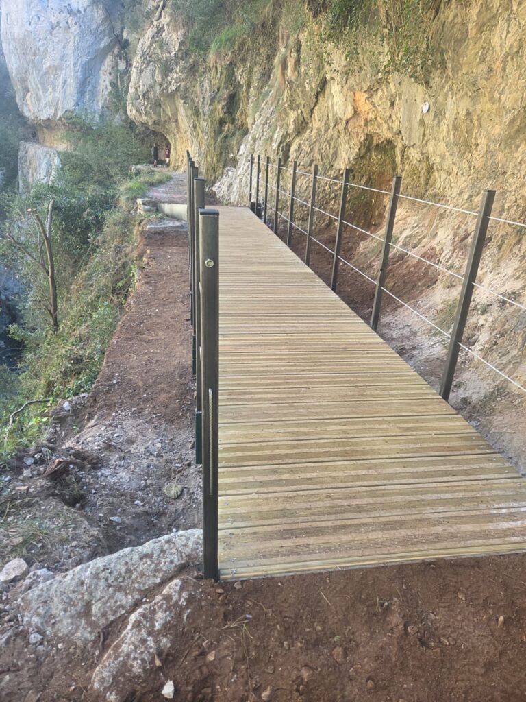 Pasarela de tablones con barandilla metálica junto a una pared rocosa.