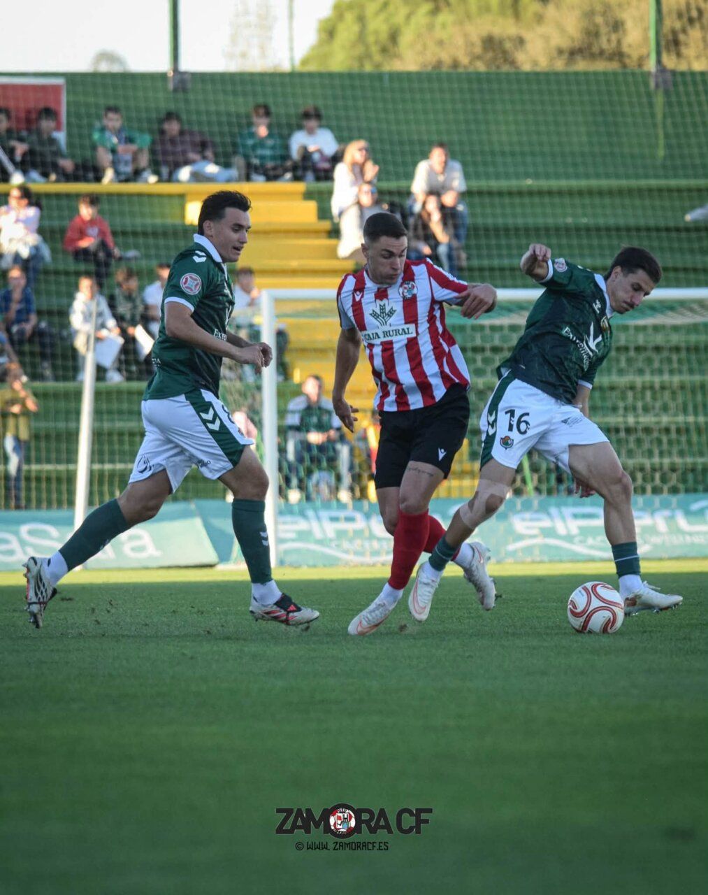 Un jugador con camiseta rojiblanca disputa el balón entre dos jugadores vestidos de verde.
