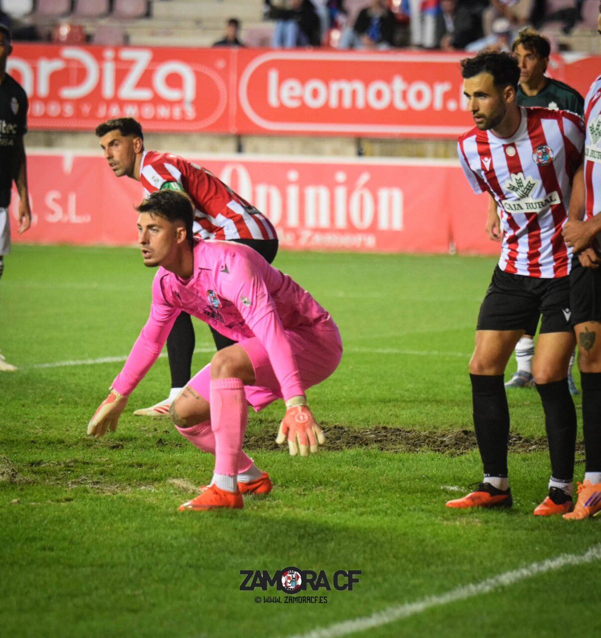 Portero de rosa agachado junto a compañeros del Zamora CF sobre el césped.
